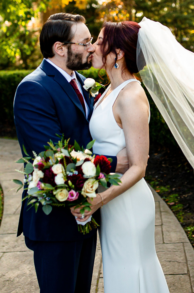 A bride in a sleeveless wedding dress and veil, holding a bouquet, kisses a groom in a suit as they stand outside on a stone path, with hedges in the background.