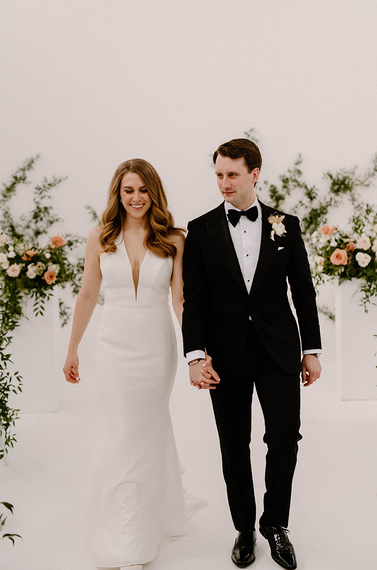A bride in a plunging neckline wedding dress and a groom in a suit hold hands and walk in a lightly decorated room.
