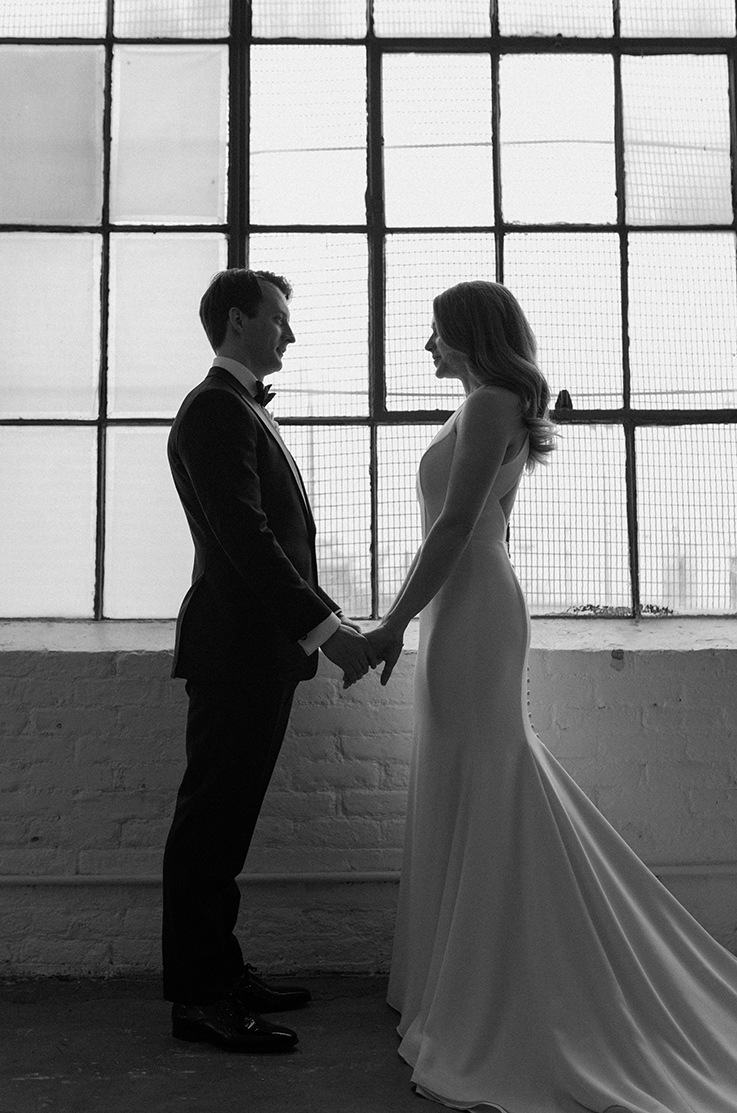 A bride in a sleeveless wedding dress and a groom in a suit face each other while holding hands and smiling, inside a brick building in front of large windows.