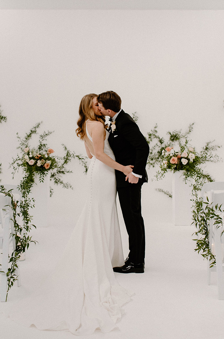 A bride in a sleeveless wedding dress and a groom in a suit kiss in a lightly decorated room.