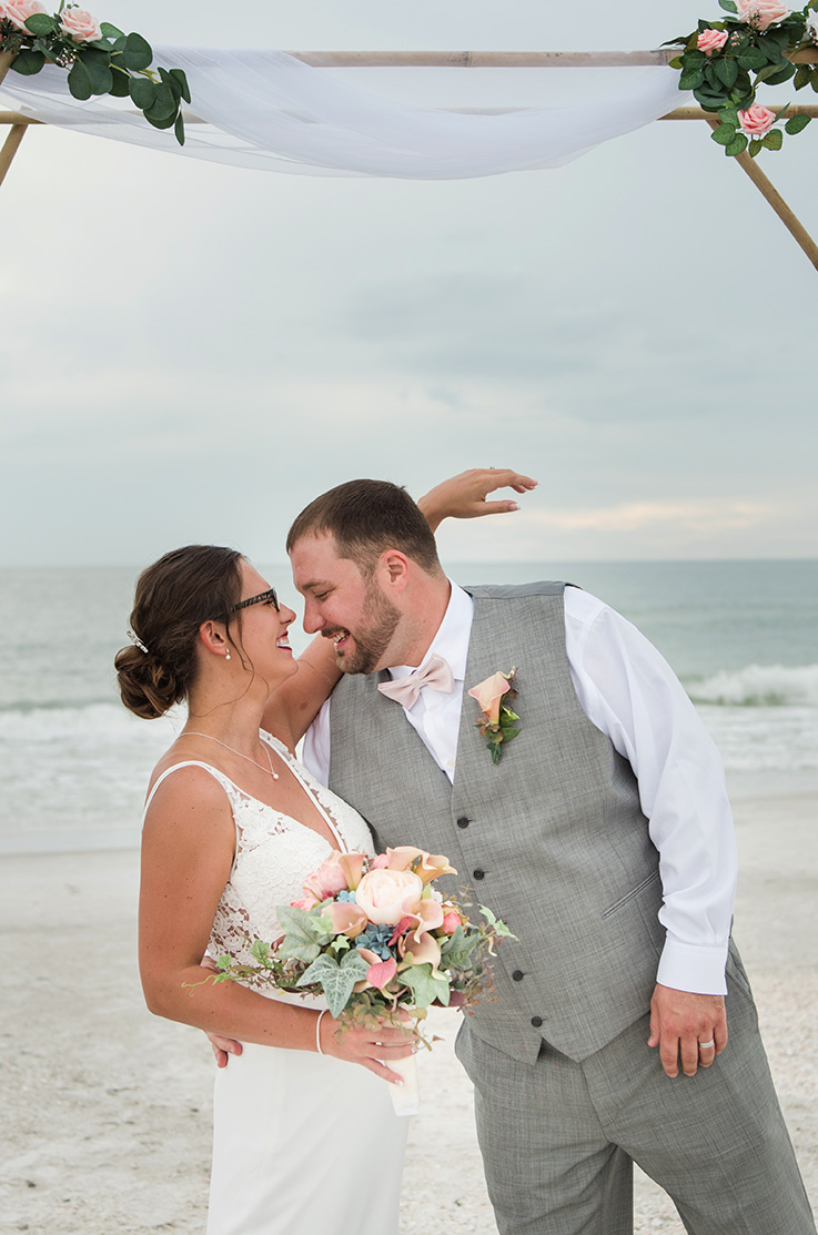 A bride in a V-neck wedding dress, holding a bouquet, and a groom in a suit vest lean in close and smile at each other, with a sandy beach and an ocean in the background.