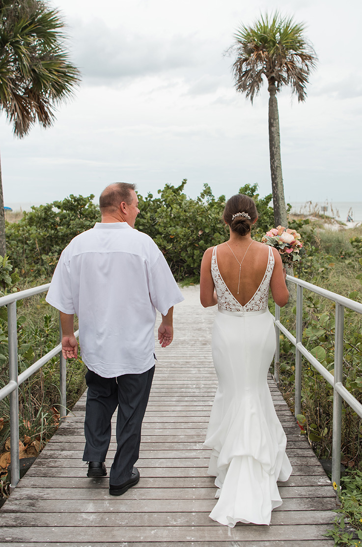 A bride in a V-back wedding dress and a groom in a short sleeve dress shirt walk across an outdoor wooden pedestrian bridge towards a path, with bushes and palm trees in the background.