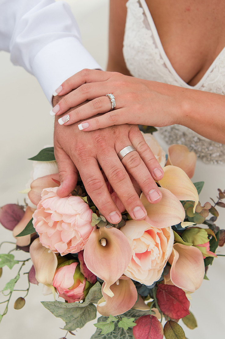 A close up of a bride's hand and a groom's hand over a bouquet of flowers, each hand has a wedding ring on it.