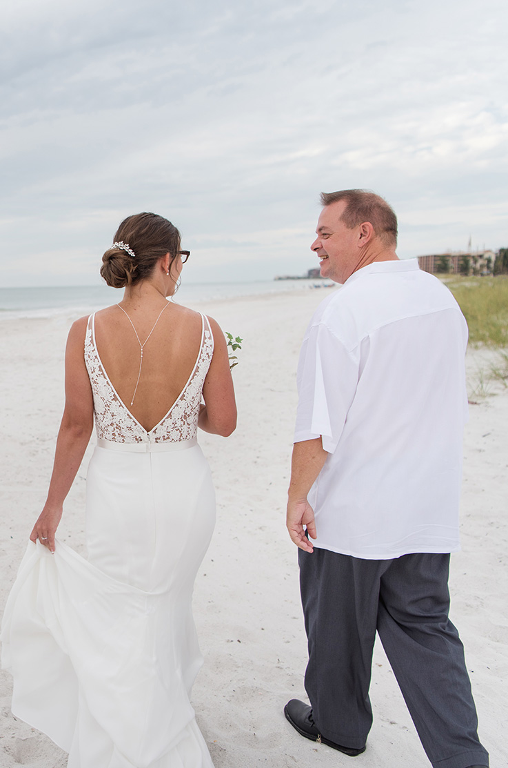 A bride in a V-back wedding dress and a groom in a short sleeve dress shirt walk along a sandy beach together.