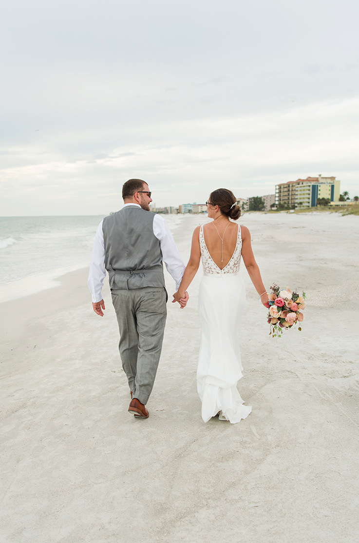 A bride in a V-back wedding dress, holding a bouquet, and a groom in a suit vest hold hands and walk down a sandy beach together.
