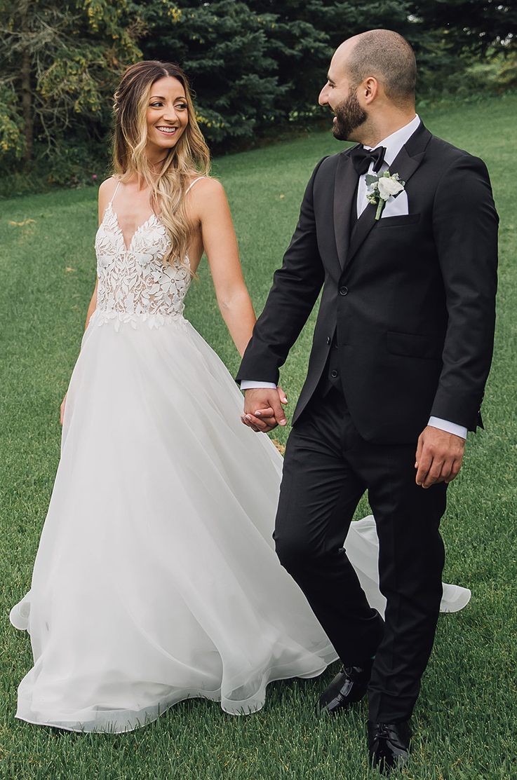 A bride in a V-neck wedding dress and a groom in a suit walk hand in hand while looking at each other smiling, outdoors in the grass with trees in the background.