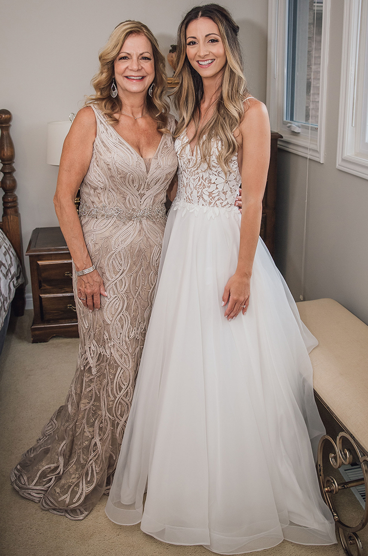 A woman in a sleeveless evening gown and a bride in a spaghetti strap wedding dress stand together and smile while standing indoors in a bedroom, with small windows behind them.