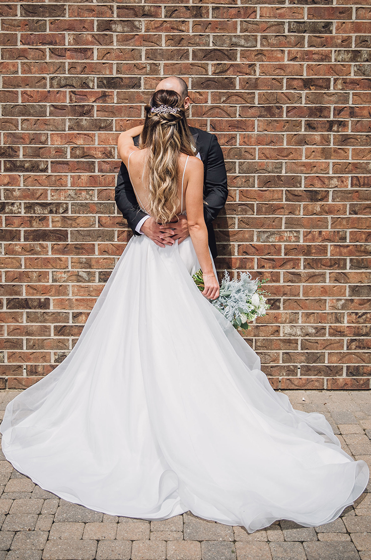 A bride in a flowing wedding dress stands in front of a groom in a suit while facing him and holding a bouquet to her side, outside in front of a brick wall. 