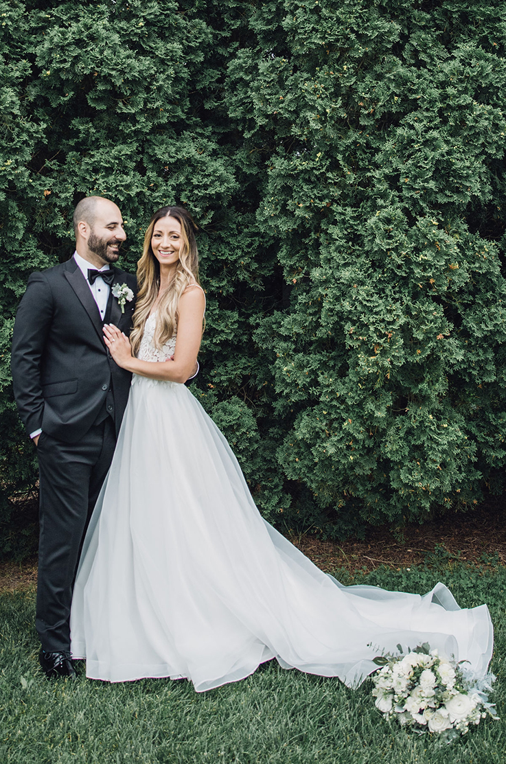 A bride in a flowing wedding dress and a groom in a suit stand together outside in the grass, in front of trees, with a bouquet at the end of the bride's train.