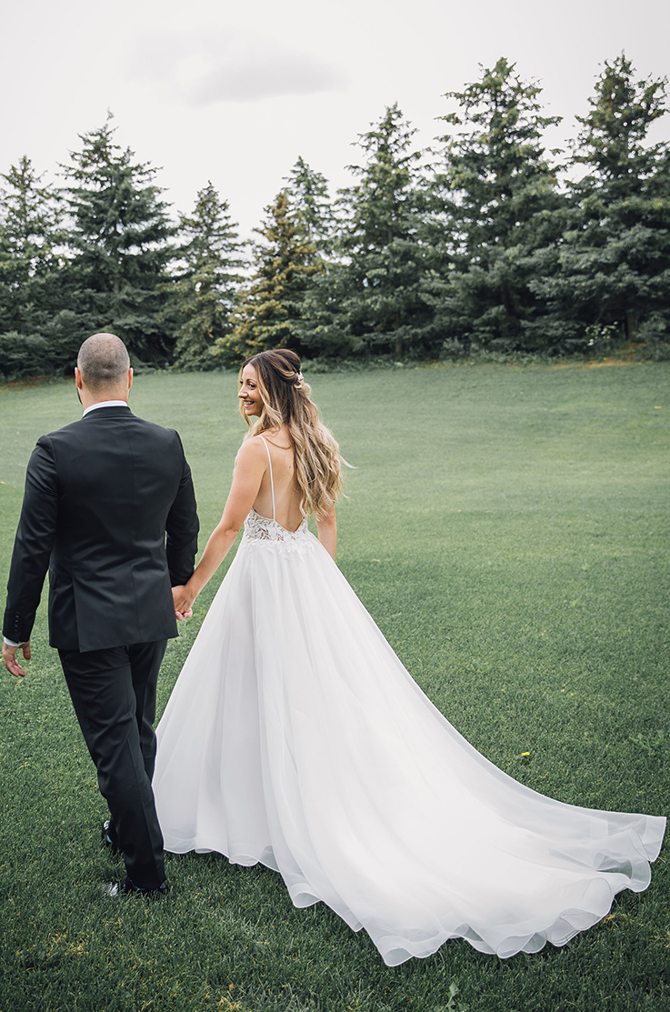 A bride in a spaghetti strap wedding dress and a groom in a suit walk together in a grassy yard, with trees in the background.