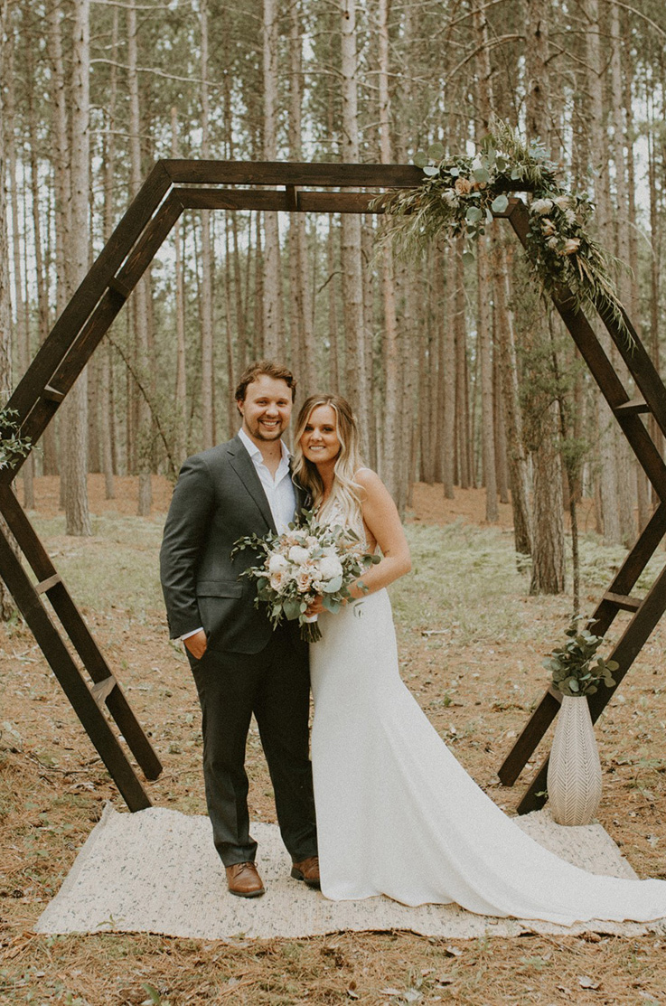 A bride in a sleeveless wedding dress, holding a bouquet, and a groom in a suit stand beside each other outdoors in a forest, under a geometric wedding arch.