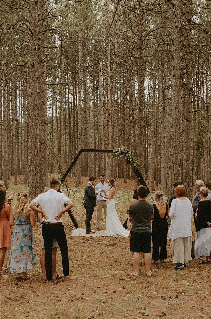 A group of wedding guests watch a bride in a sleeveless wedding dress and a groom in a suit exchange vows in a forest, under a geometric wedding arch.