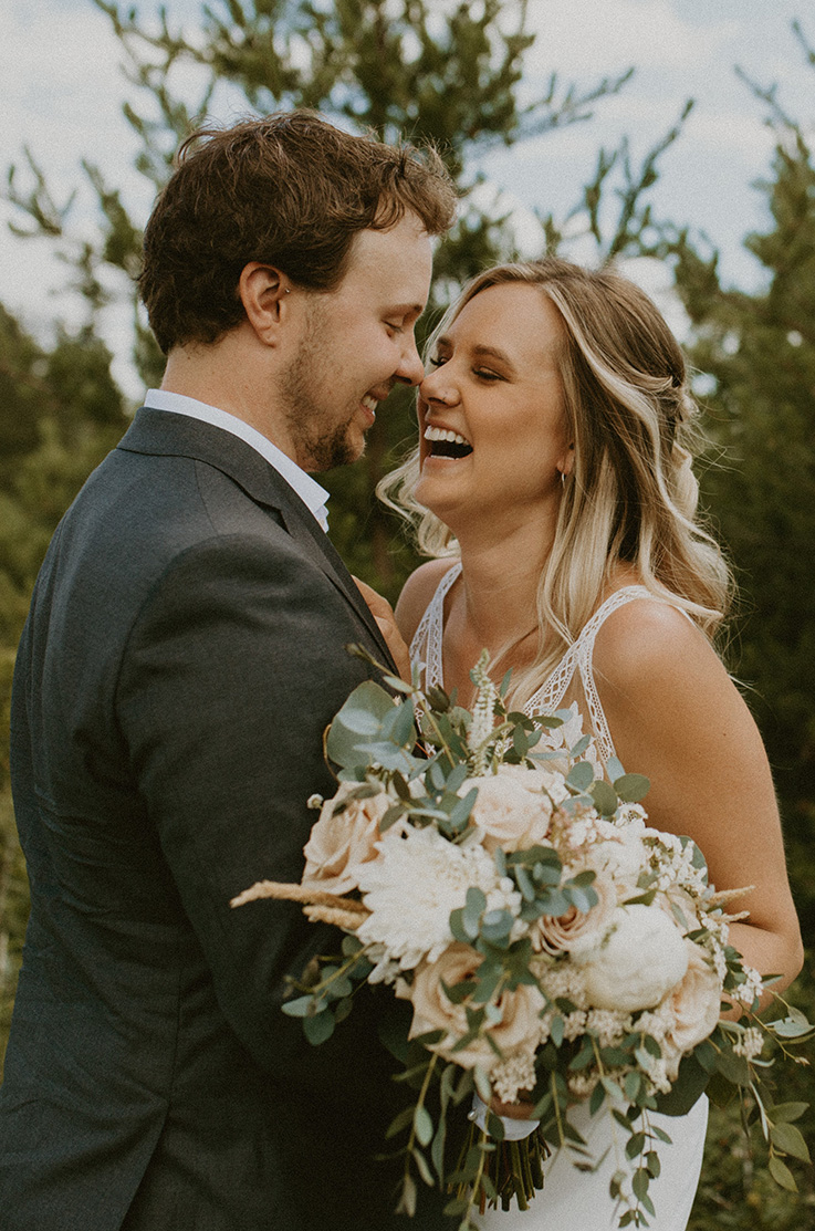 A bride in a sleeveless wedding dress, holding a bouquet, and a groom in a suit hold each other and laugh while standing outdoors, with trees in the background. 