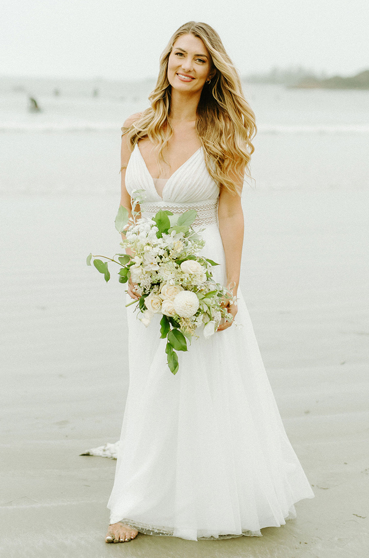 A bride in a V-neck wedding dress smiles and holds a bouquet while standing on a sandy beach.