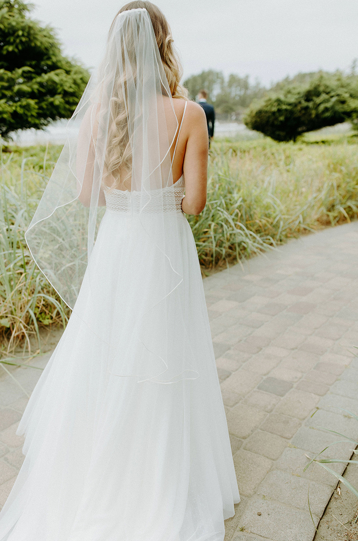 A bride in a tulle wedding dress and veil walks down a stone path outdoors, surrounded by tall grass with trees in the background.