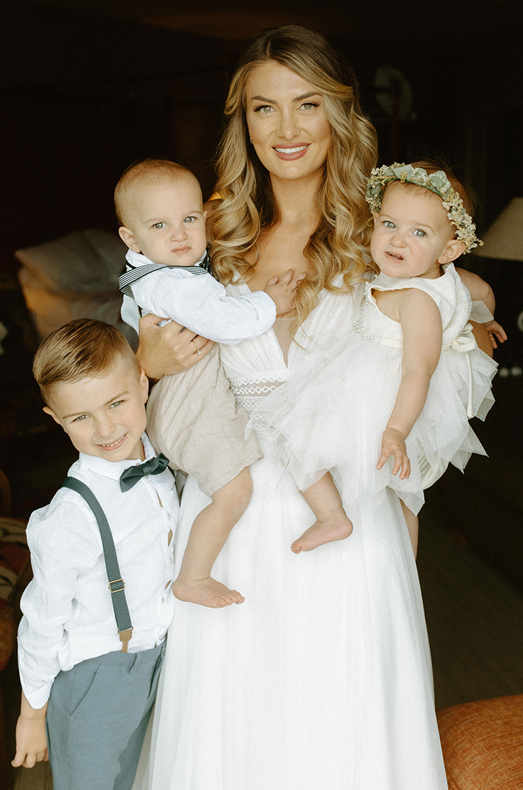 A smiling bride in a tulle wedding dress holds two babies, one in each arm, and a small boy in suspenders smiles and stands by her side, while they stand indoors.