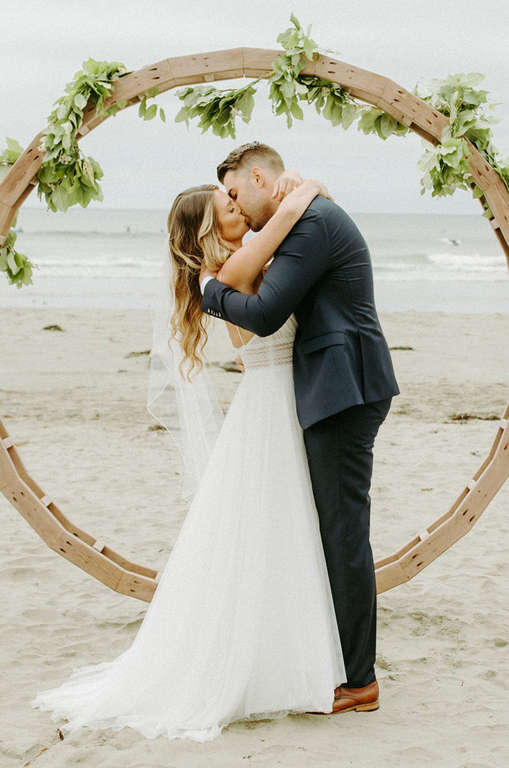 A bride in a tulle wedding dress and a groom in a suit kiss in front of a circular wedding decoration on a sandy beach, with an ocean in the background.