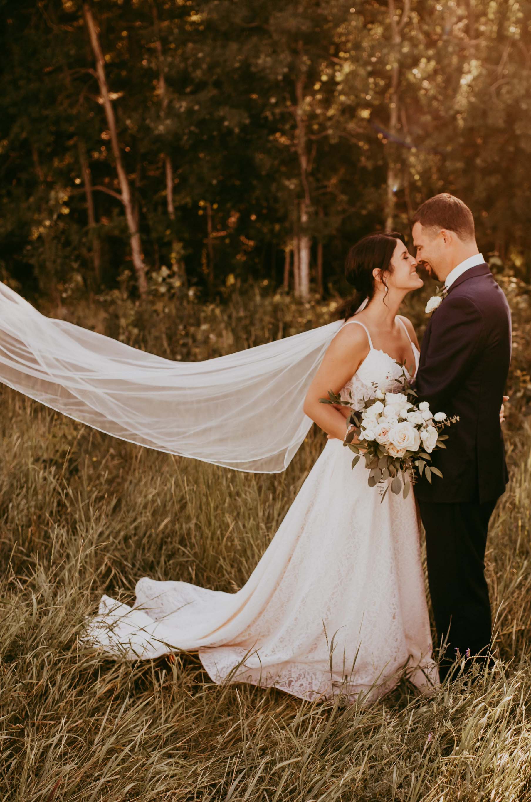 Groom embraces bride in a lace sweetheart neck wedding dress with a flowing veil, and is holding a bouquet in forest meadow.