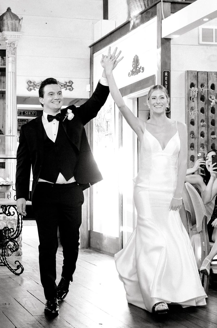 Bride in a Mikado spaghetti strap gown with a V-neck and princess seams, enters venue with her groom in black and white.