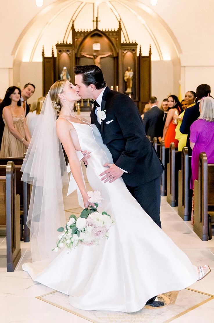 Groom dips his bride in a V-neck Mikado wedding dress with a veil and is holding a bouquet, as they kiss in a church aisle.