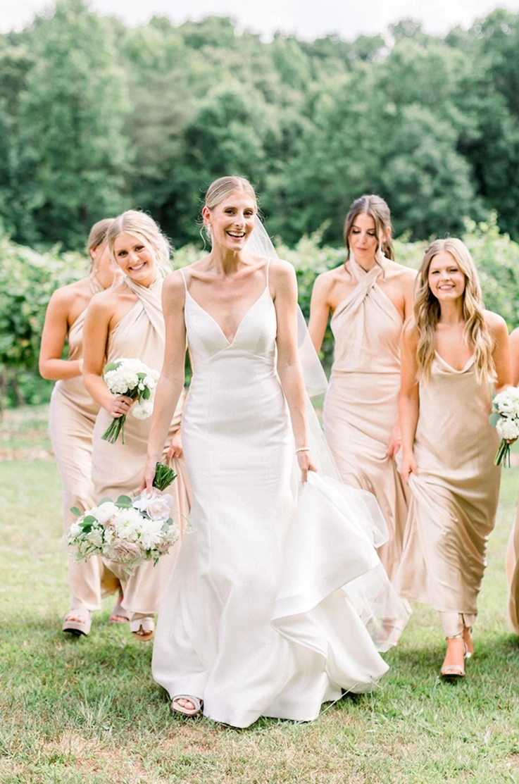 Bride in a V-neck empire waist gown in Mikado, holds her train and a bouquet among bridesmaids in satin dresses outdoors.