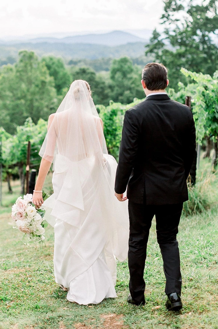 Bride in a low back Mikado gown with a bow and a veil, holds her train and walks alongside the groom down a vineyard.