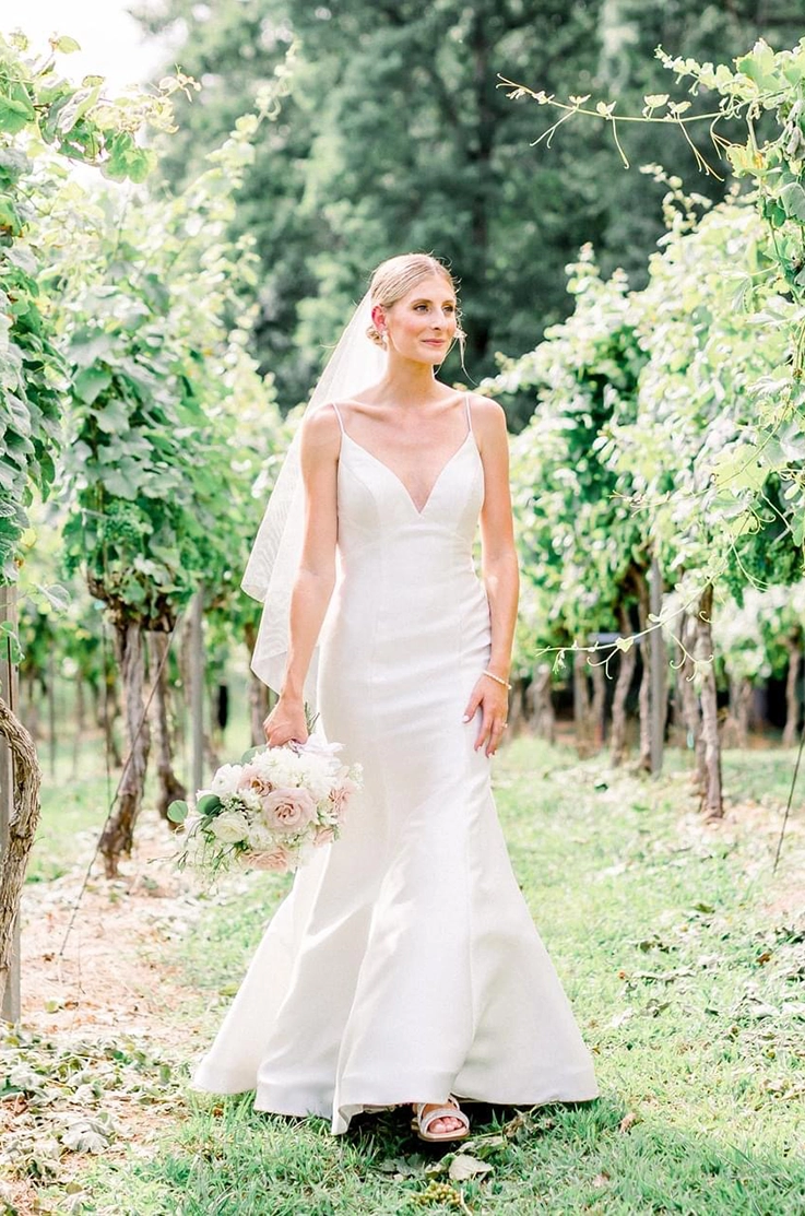 Bride poses in a vineyard row, in a fit and flare Mikado gown with a V-neck and princess seams, holding a blush bouquet.