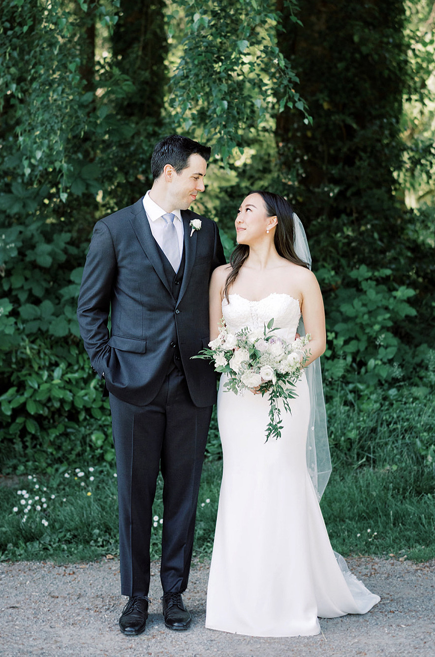 Bride in a strapless crepe gown with lace detail, a fit and flare skirt and a veil, looks up at her groom on a gravel path.