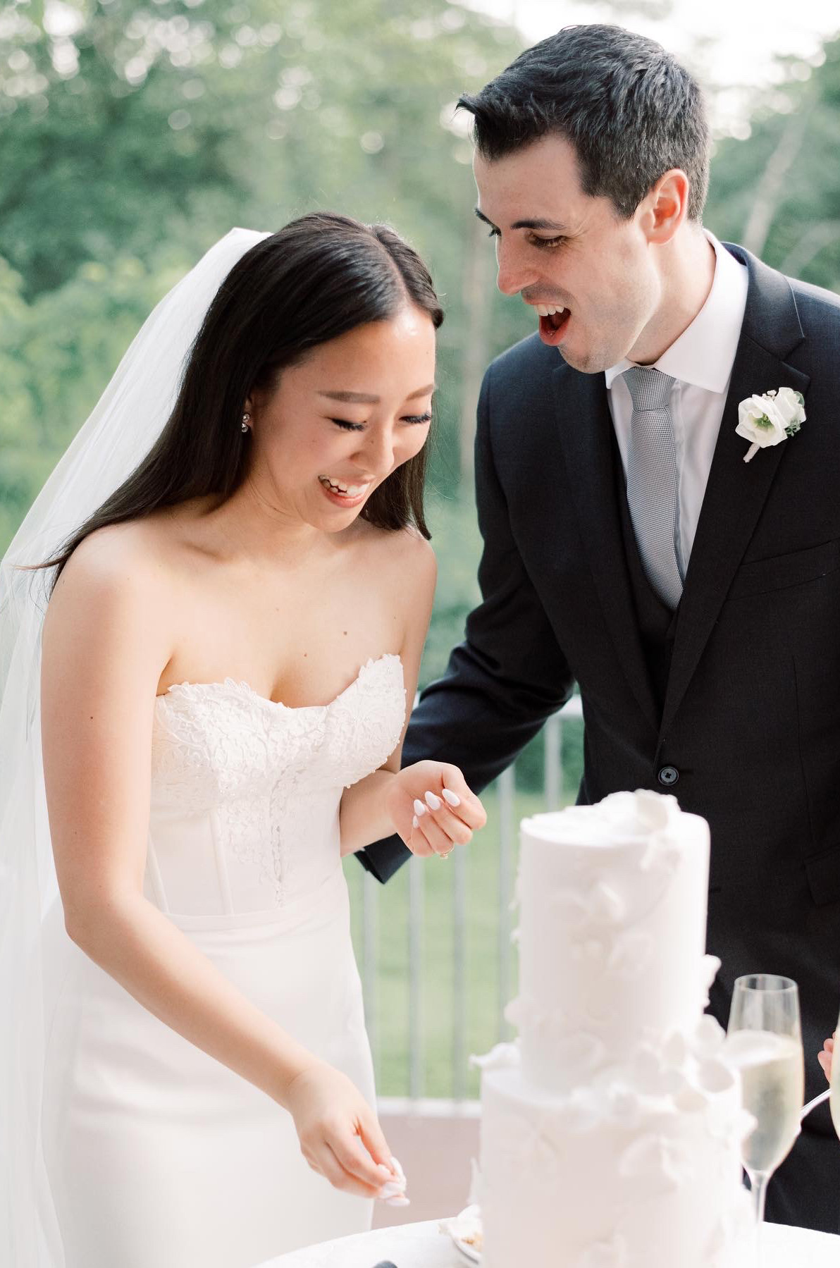 Bride in a strapless crepe wedding dress with lace detail and a veil, smiles cutting cake next to the groom outdoors. 