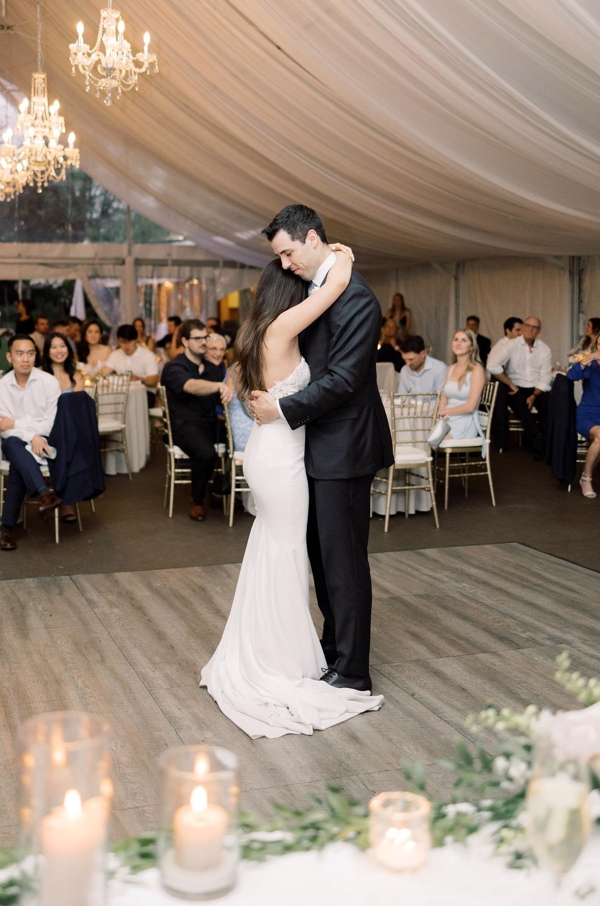 Groom dances with bride in a strapless fit and flare gown with lace appliques under draped curtains and chandeliers.