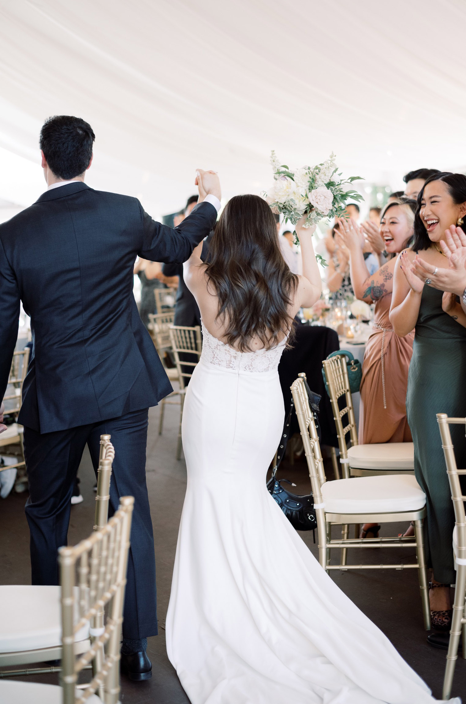 Bride in a strapless crepe wedding dress with a lace back and fit and flare skirt, celebrates with groom in a tented reception.