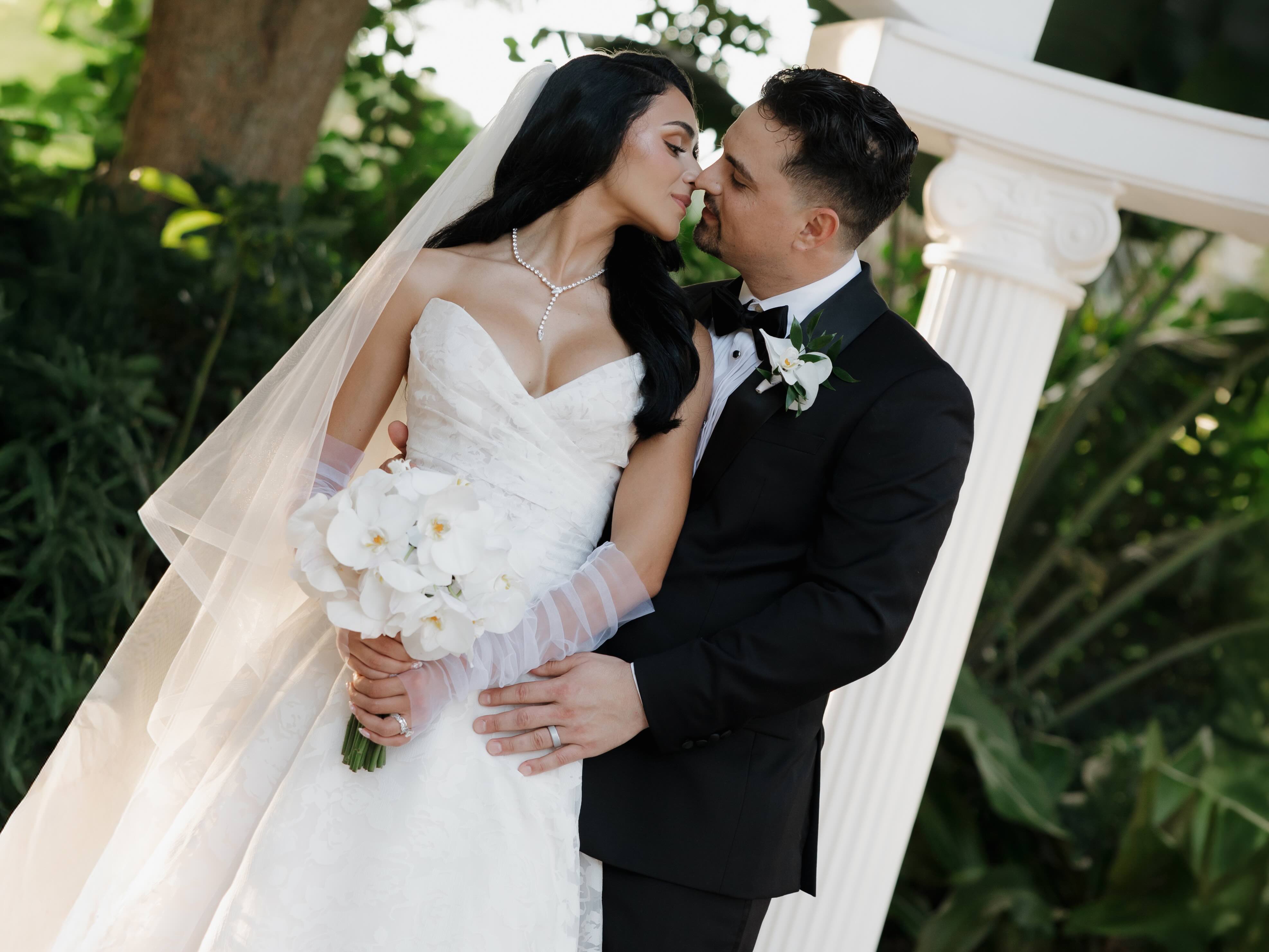 A bride in a fitted lace wedding gown embraces her groom beneath a white column archway surrounded by greenery, holding a bouquet of white orchids.