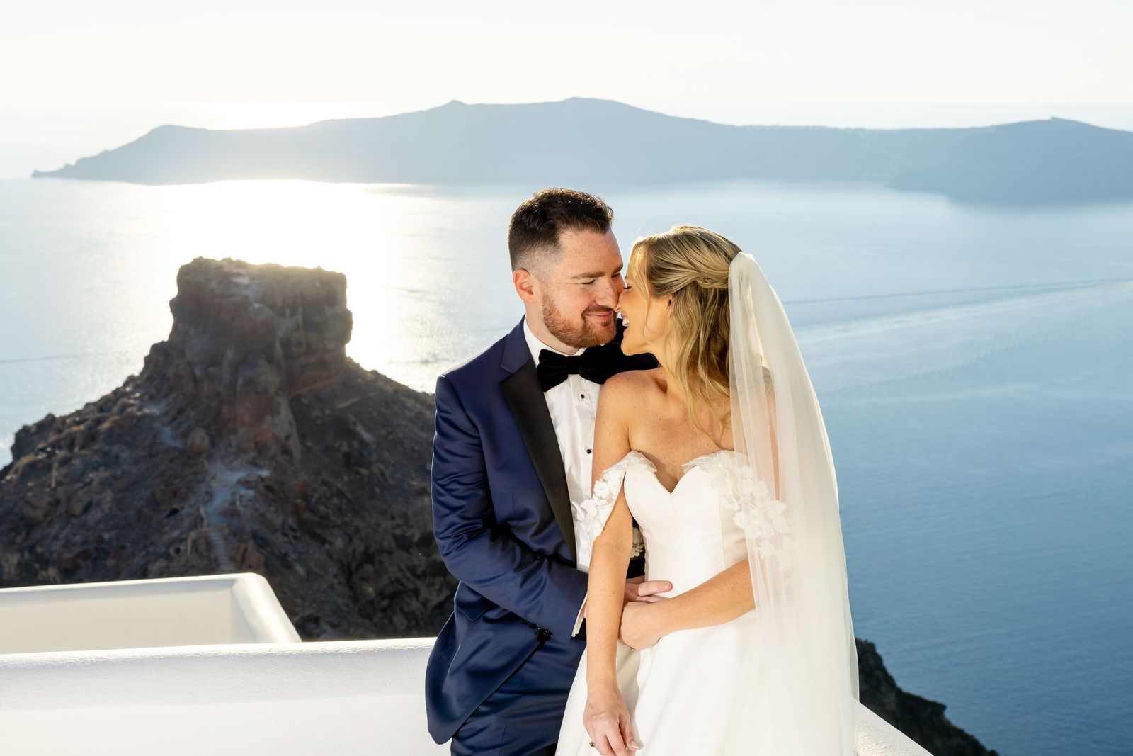 A bride in an off-the-shoulder wedding gown with floral lace sleeve detail shares a kiss with her groom on a cliffside terrace overlooking the sea.