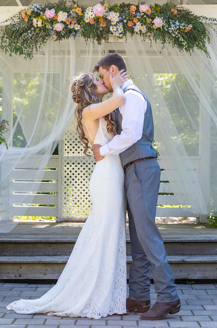 Groom kisses his bride in a lace fit and flare gown with a low open back, under a white floral arbor with curtains.