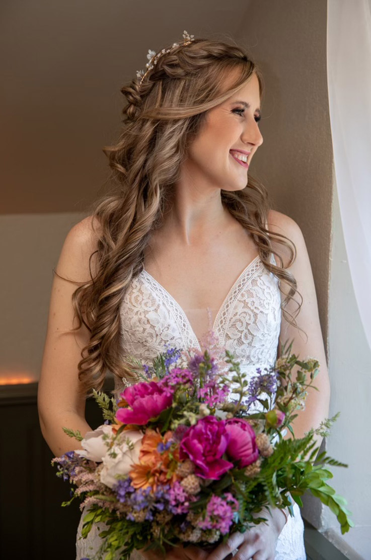 Close-up of a bride looking out a window in a sleeveless lace gown with a plunging V-neck, holding a colorful bouquet.