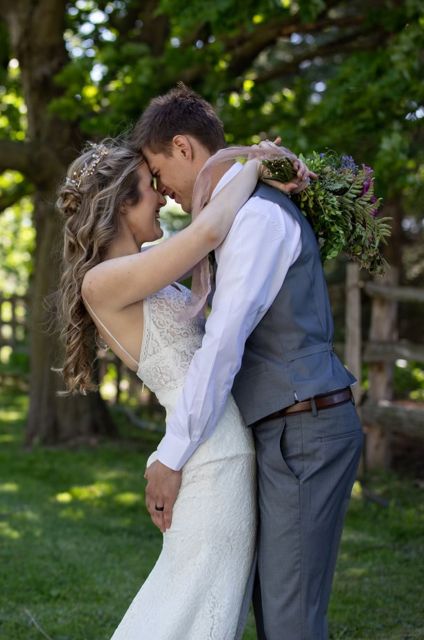Groom faces his bride in a sheer lace bodice wedding dress with thin straps, and is holding a bouquet in a sunny outdoor area.