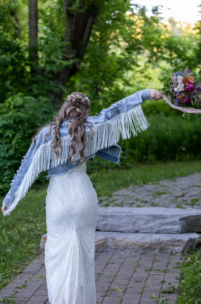 Back of bride in a fit and flare lace gown with a denim fringe jacket overtop, posing with a bouquet on a stone garden path.