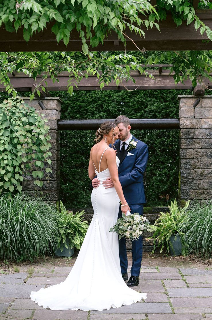 Groom embraces his bride in a low back crepe wedding dress with a buttoned fit and flare skirt, holding a bouquet in a garden.