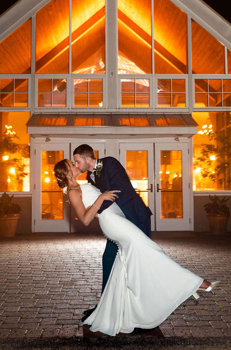 Groom dips his bride in a pleated crepe bodice gown with a fit and flare skirt, and they kiss in front of warmly lit venue.