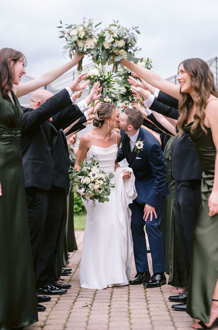 Groom kisses his bride holding her train in a scoop neck crepe gown, crouching under guests raising their bouquets.