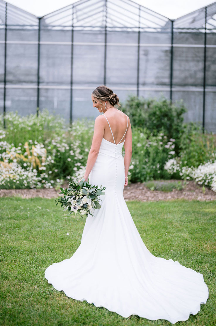 Back of bride in a pleated crepe bodice gown with a low back, buttons and a fit and flare skirt, holding a bouquet outdoors.