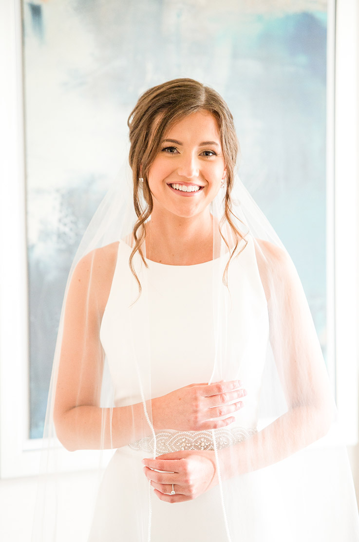 Close-up of bride in a sleeveless crepe gown with a bateau neck, belt and a veil, stands by a painting in a brightly lit room.
