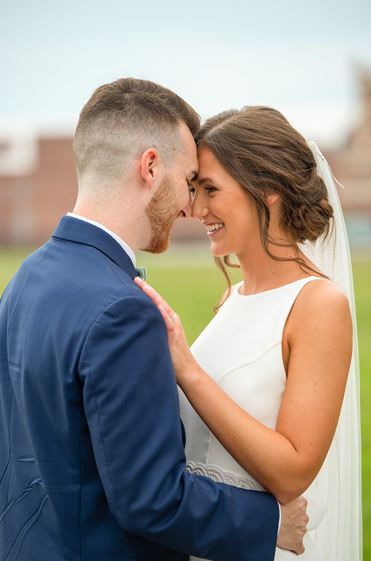 Groom holds bride in an embrace in a bateau neck crepe gown with belt detail and a veil, facing each other outdoors.