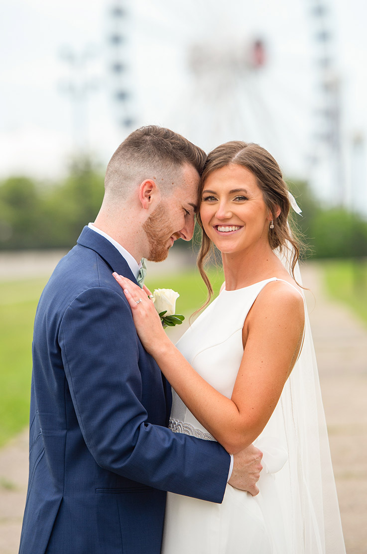 Groom embraces his bride in a sleeveless crepe gown with a high neckline, belt and a veil, posing on an outdoor path.