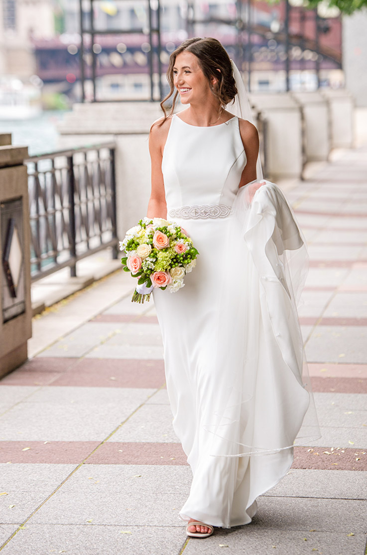 Bride in a bateau neck crepe wedding dress with a beaded belt and a veil, holds her train smiling on a waterfront walkway.