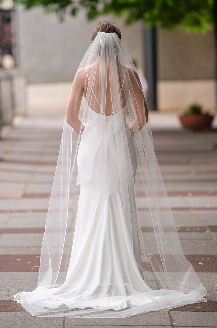 Back of bride in an open back crepe gown with a sash, thin straps and a long veil, is walking on a brick pathway.