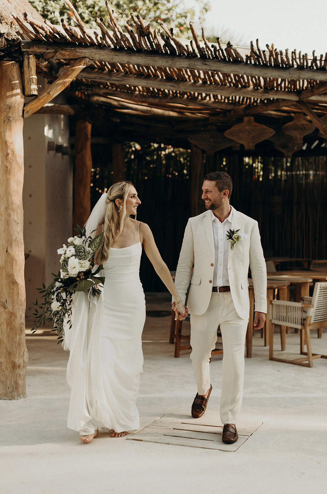 Bride in a fit and flare crepe wedding dress with pleating and a veil, holds her train and walks hand in hand with the groom. 