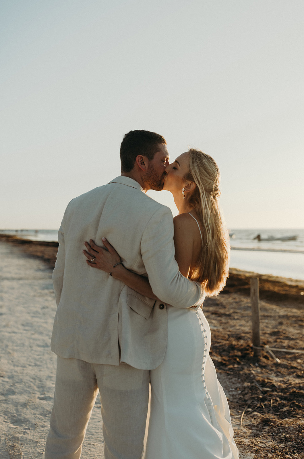 Groom kisses his bride in a sleeveless crepe fitted gown with buttons and a low back, as they stand on a beach shore.