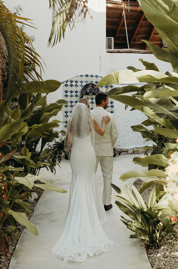 Bride walks in a fit and flare crepe gown with a low back and a veil, behind groom on a pathway surrounded by tropical plants.
