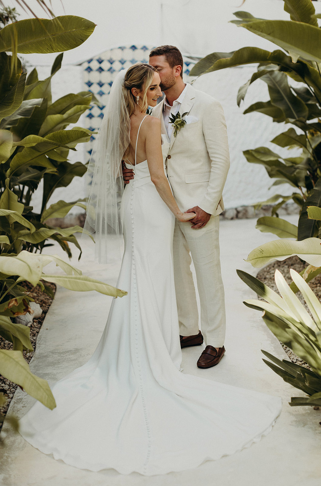 Groom embraces bride in an open back crepe gown with a pleated bodice, fit and flare skirt and a veil, in a tropical courtyard.